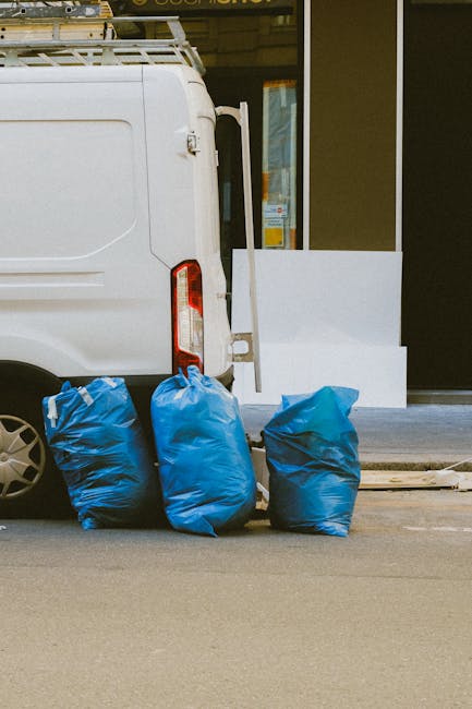 A white commercial van parked on a street outside a building with large glass windows. The rear doors of the van are closed, and on the pavement next to it are three large blue recycling bags filled with household waste, likely including furniture or non-recyclable materials. Behind the van, a portion of the building's entrance is visible, with a white step and part of the door frame. The scene depicts elements typical of home relocation or waste disposal services, with the van possibly used by Man With a Van Borough for furniture transport or bulky waste removals as part of a house removal process. The overall setting suggests preparation for collection or disposal, emphasizing logistics involved in furniture transport and packing during a move or clearance.