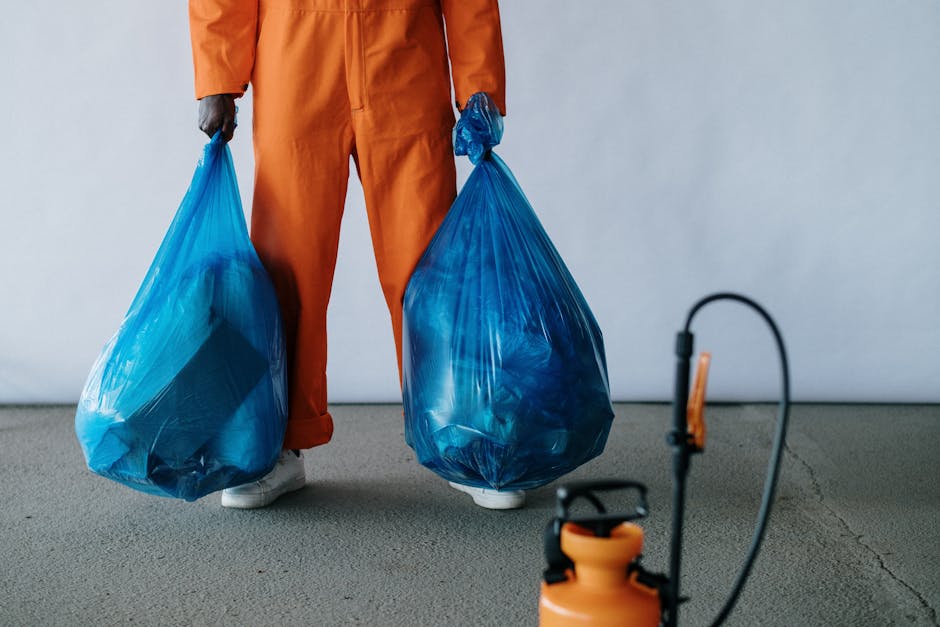 A person wearing orange work overalls and white shoes is standing indoors on a gray concrete floor, holding two large blue rubbish bags filled with waste material, suggesting preparation for bulky waste or furniture disposal as part of a home relocation or moving process. The background features a plain white wall, indicating an indoor setting such as a garage or storage area. Nearby, a black and orange sprayer or cleaning device is visible, possibly used for cleaning or pest control during move-in or move-out activities. The person's face and upper body are not shown, focusing on the action of handling waste bags which are positioned at waist height. This scene illustrates the logistics involved in furniture disposal or clearance, a step often associated with professional removals services offered by companies such as Man With a Van Borough, supporting house removals, packing, and transport logistics.