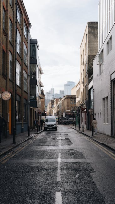 A narrow urban street scene showing the exterior of multi-story buildings on both sides, with a white van parked in the middle of the road. The pavement appears wet, reflecting the surroundings, and a few pedestrians are visible on the sidewalk. In the background, taller modern buildings and a cloudy sky can be seen. The image depicts a typical city environment suitable for house removals and furniture transport operations. Occasionally, Man With a Van Borough's services for home relocation and packing logistics are relevant, especially during the loading and unloading process of items like boxes, furniture, or appliances. The street's limited access and urban setting highlight the importance of tight-access van solutions provided by professional removals companies in the Borough area.