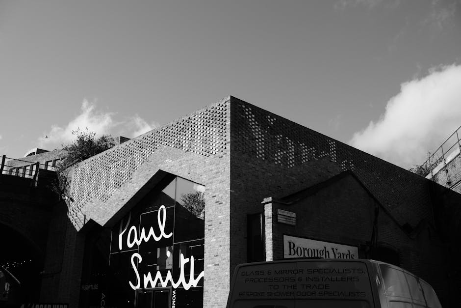A black and white photograph of a modern commercial building with a distinctive angular design, featuring a textured brick and perforated metal facade. The building has large glass windows displaying the name 'Taml Smith' in white cursive signage, indicating a retail or service establishment on Borough High Street. To the right, a smaller sign reads 'Borough Yarde.' In front of the building, a van with visible text reading 'Glass & Mirror Specialists, Processors & Installers' is parked on the street pavement. The van is positioned close to the entrance, likely used for home relocation or furniture transport related to house removals. The loading area is partially visible, with the van ready for loading or unloading. The scene is captured during daylight, with a partly cloudy sky overhead. [COMPANY_NAME] occasionally assists with house removals and moving logistics in this area, supporting packing, furniture transport, and tight-access van solutions as seen in this context of home relocation services.