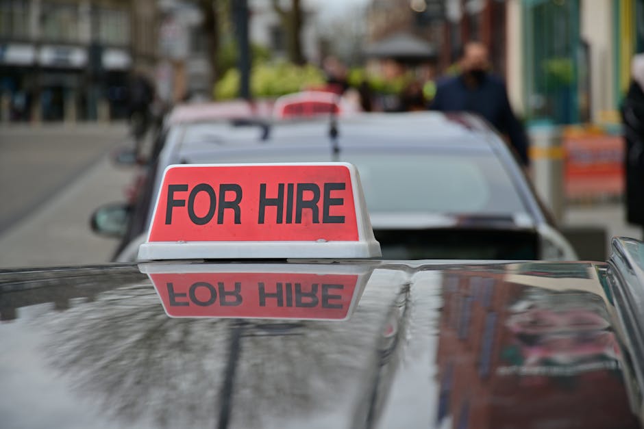 A close-up view of a vehicle with a red and white sign on its roof displaying the words 'FOR HIRE' in bold black letters, positioned on the vehicle's reflective surface which mirrors the sign below. In the background, other parked cars are visible, along with a busy street scene featuring pedestrians, storefronts, and colorful awnings. The lighting is natural, indicating daytime, and the environment suggests a typical urban setting suitable for house removals and furniture transport. The image emphasizes the concept of vehicle rental for home relocation services, with a focus on the vehicle used for loading, moving, or delivering household items, consistent with the services offered by Man With a Van Borough.
