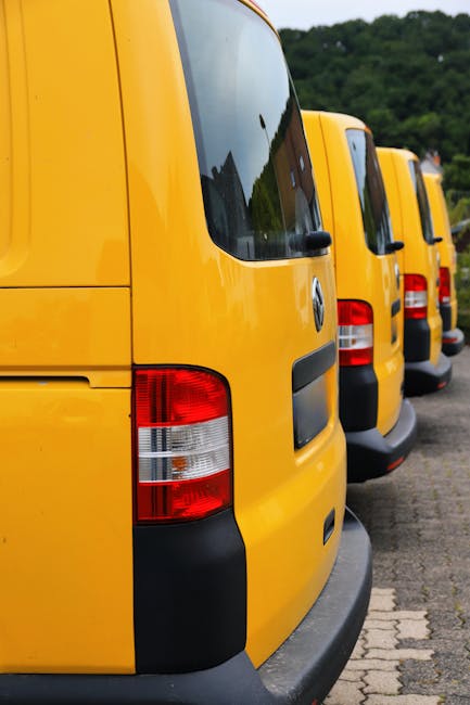 A lineup of four yellow commercial vans parked side by side on a paved surface, with their rear doors facing the viewer. Each van has a black bumper and red-and-white tail lights, and all are clean and in good condition. The vans are part of a house removal or furniture transport service, possibly associated with Man With a Van Borough, and are arranged in a straight formation next to a curb. The background shows a green, tree-lined area indicating an outdoor setting, with natural daylight illuminating the scene. This image represents the loading process involved in home relocation, involving vehicle transport for furniture and boxes, with the vans ready to assist in packing, moving, or delivery tasks typical of professional removals services.