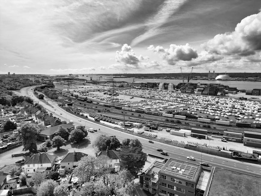 A black-and-white aerial image showing an expansive industrial port area with numerous shipping containers and cranes situated along the waterfront, extending into the distance under a partly cloudy sky. In the foreground, a residential neighbourhood with houses, trees, and a main road featuring moving vehicles is visible. The port is bordered by a railway line and the scene includes a large parking lot filled with vehicles, suggesting a busy logistics hub. The illustration of the port and surrounding infrastructure reflects aspects of home relocation and furniture transport logistics, as part of the process of packing and loading in house removals. The overall scene captured by Man With a Van Borough emphasizes the scale of commercial transport and the urban environment involved in moving and relocation services.