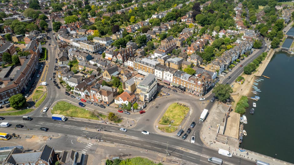 An aerial view of a town centre featuring a mix of residential and commercial buildings with pitched roofs, situated next to a waterway with several moored boats. In the foreground, a busy multi-lane road with moving cars, vans, and trucks runs parallel to the water, while a small roundabout connects to smaller streets leading into the town. To the right, an open loading area near the water contains parked vehicles and a few stacked cardboard boxes, plastic wrapping, and packing materials, suggesting ongoing home relocation or furniture transport activities. The scene is well-lit with natural daylight, highlighting the urban layout, trees, and surrounding green spaces, providing a comprehensive visual context for house removals and moving services such as those offered by Man With a Van Borough.