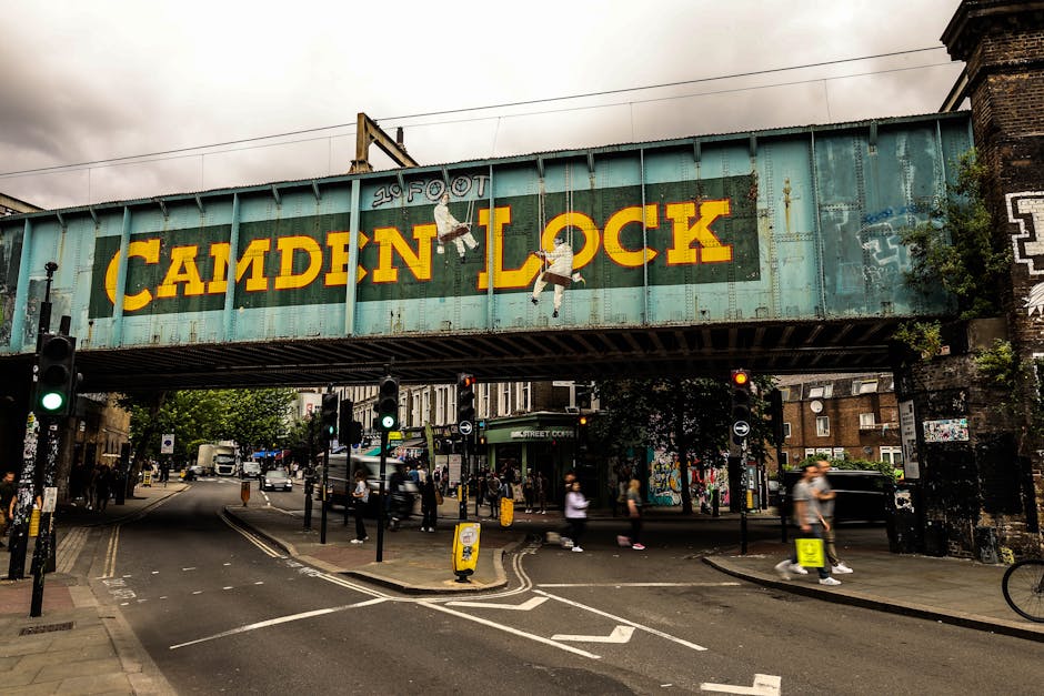 A large, weathered railway bridge spanning above a busy street in Camden, London, featuring a prominent, brightly painted sign reading 'Camden Lock' in yellow letters on a blue-green background. The bridge's surface shows signs of rust and aging, with a mural of children climbing on the lettering. Below, the intersection includes pedestrian crossings with zebra stripes, traffic signals showing green and red lights, and several pedestrians walking on sidewalks or crossing the street. A few cyclists are also visible, some waiting to cross. On the street level, there are parked cars and delivery vans, along with small street-level shops and street art on the building walls. The scene is captured during cloudy daytime, with urban environment details such as street signs, lampposts, and some greenery near the buildings, reflecting the vibrant, dynamic atmosphere typical of home relocation or furniture transport logistics in a city setting, as observed by [COMPANY_NAME].