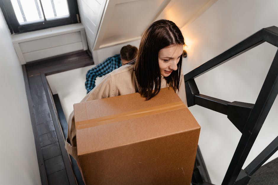 A woman with shoulder-length dark hair, wearing a beige coat, is carrying a large cardboard box during a house removal inside a residential property. She is walking down a staircase with a black metal railing, with a man in a blue checkered shirt assisting behind her. The staircase is narrow and enclosed, with a stair tread visible beneath her and a wooden door at the top. Inside the property, the walls are painted white, and there is a warm light source illuminating the scene. The cardboard box is sealed with packing tape and appears to contain household items for a home relocation. Behind her, the man is partially visible, also holding a box, suggesting a coordinated effort in furniture transport and packing during the moving process. This image demonstrates careful handling of belongings, typical of professional removals carried out by companies like Man With a Van Borough, specializing in safe and efficient moving services in Borough.