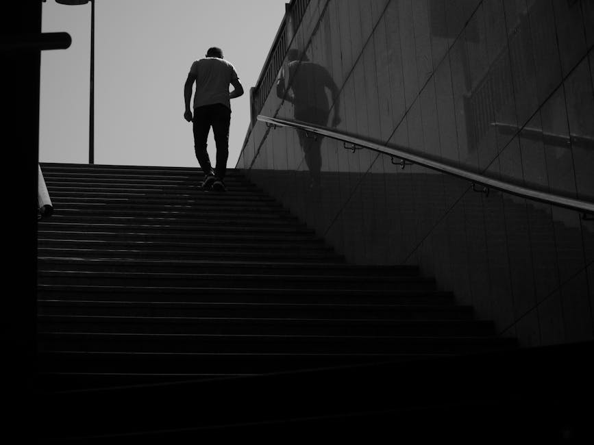 A person is seen from below as they ascend a wide outdoor staircase with dark steps, carrying a bag over their right shoulder. The individual is dressed in casual clothing and is framed against a clear, light sky. To the right side, a modern building's reflective glass facade mirrors the person's image, creating a subtle reflection effect. The staircase is adjacent to the building, which features clean, smooth panels. On the left, part of a vehicle is visible, indicating a possible loading or moving area. The scene is illuminated by natural daylight, with the emphasis on the person's movement and the urban environment. This image, associated with home relocation services, emphasizes the process of transporting belongings up stairs as part of a secure and professional moving or removal operation, such as those offered by Man With a Van Borough.