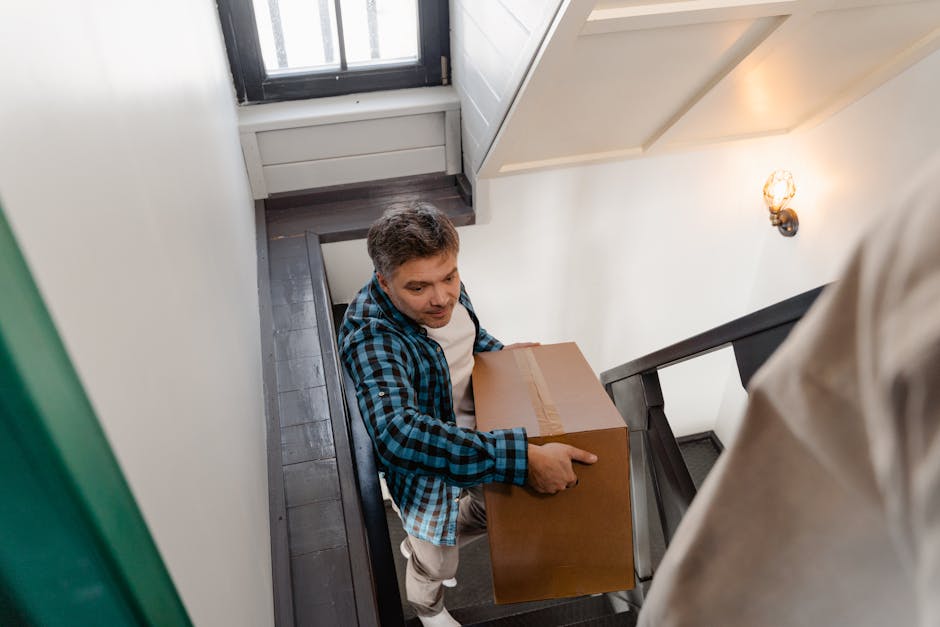 A woman with shoulder-length dark hair, wearing a beige coat, is carrying a large cardboard box during a house removal inside a residential property. She is walking down a staircase with a black metal railing, with a man in a blue checkered shirt assisting behind her. The staircase is narrow and enclosed, with a stair tread visible beneath her and a wooden door at the top. Inside the property, the walls are painted white, and there is a warm light source illuminating the scene. The cardboard box is sealed with packing tape and appears to contain household items for a home relocation. Behind her, the man is partially visible, also holding a box, suggesting a coordinated effort in furniture transport and packing during the moving process. This image demonstrates careful handling of belongings, typical of professional removals carried out by companies like Man With a Van Borough, specializing in safe and efficient moving services in Borough.
