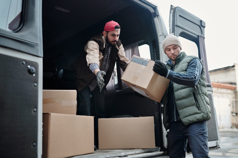 Two men are loading cardboard boxes into the rear of a dark grey van parked outside a building. One man, wearing a red baseball cap, a beige jacket with rolled-up sleeves, and grey gloves, is leaning into the van to place a box on top of other packed boxes inside. The second man, dressed in a grey beanie, a grey quilted vest over a blue hoodie, and black gloves, is standing on the pavement, holding a box and preparing to load it into the vehicle. The scene is outdoors during daytime, with the van’s open rear door revealing the interior filled with packed cardboard boxes, some sealed with packing tape. The packaging materials include cardboard, and the van is equipped with a trolley and securing straps visible near the entrance. This image reflects the process of home relocation, furniture transport, or packing and moving logistics, with the men coordinating to facilitate a smooth loading process for a house removal, as part of professional services offered by Man With a Van Borough.