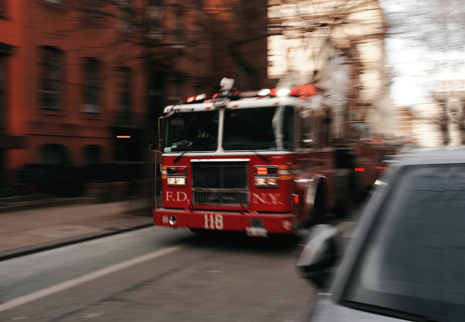 A red fire engine with emergency lights on the roof is parked on a city street in front of a multi-story brick building, slightly blurred due to motion. The vehicle features black vents and white lettering on the front, indicating it belongs to the Fire Department. Nearby, a black or dark grey vehicle is partly visible in the foreground, parked close to the curb. The scene captures an urban environment during daylight, illustrating a situation where emergency services might be responding during a home relocation or moving process. Man With a Van Borough's removals service is applicable in such scenarios, involving fast transport, loading, and handling of household furniture and boxes, often requiring quick or emergency move arrangements.