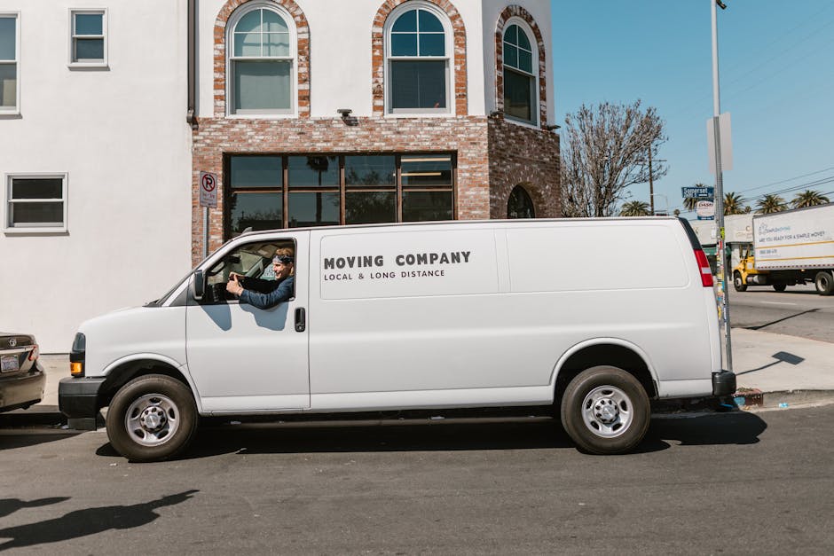 Two men are loading cardboard boxes into the rear of a dark grey van parked outside a building. One man, wearing a red baseball cap, a beige jacket with rolled-up sleeves, and grey gloves, is leaning into the van to place a box on top of other packed boxes inside. The second man, dressed in a grey beanie, a grey quilted vest over a blue hoodie, and black gloves, is standing on the pavement, holding a box and preparing to load it into the vehicle. The scene is outdoors during daytime, with the van’s open rear door revealing the interior filled with packed cardboard boxes, some sealed with packing tape. The packaging materials include cardboard, and the van is equipped with a trolley and securing straps visible near the entrance. This image reflects the process of home relocation, furniture transport, or packing and moving logistics, with the men coordinating to facilitate a smooth loading process for a house removal, as part of professional services offered by Man With a Van Borough.
