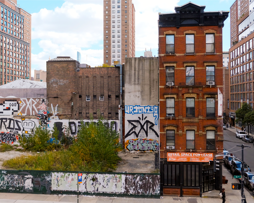 A cityscape viewed from a high angle showing a mix of old and modern buildings along Borough High Street, with a small plot of overgrown land and graffiti-covered walls in the foreground. The plot contains green bushes and weeds, and is partially enclosed by a metal fence with peeling paint. To the right, there is a narrow brick building with five floors, featuring large windows with black frames, some with air conditioning units, and a sign at street level advertising retail space for lease. The street below has parked cars and a pedestrian walking on the sidewalk. In the background, tall modern office buildings are visible under a partly cloudy sky. This scene captures the urban environment relevant to house removals and transportation logistics as referenced in the 'Parking suspensions on Borough High Street' guide, with visible elements suggesting potential obstacles and considerations for home relocation or furniture transport activities, supported by the professional services of Man With a Van Borough.