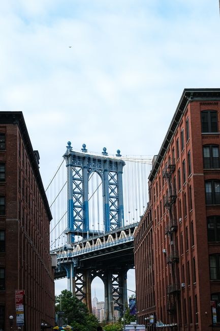 A clear daytime view of the Manhattan Bridge seen from the street level between two red brick buildings in Borough, London. The bridge's steel structure, painted blue, features suspension cables and towers with decorative finials. In the foreground, the pavement is visible with construction or moving equipment, including a trolley and plastic-wrapped packing materials, indicating active moving or packing preparations aligned with house relocation services. The architecture of the surrounding buildings shows traditional brick facades with multiple windows and fire escapes. Overhead, the sky is partly cloudy, and the overall scene reflects urban transport logistics related to furniture transport and moving processes, with the famous bridge serving as a backdrop for logistical activities supported by companies like Man With a Van Borough, specializing in removals and home relocations.