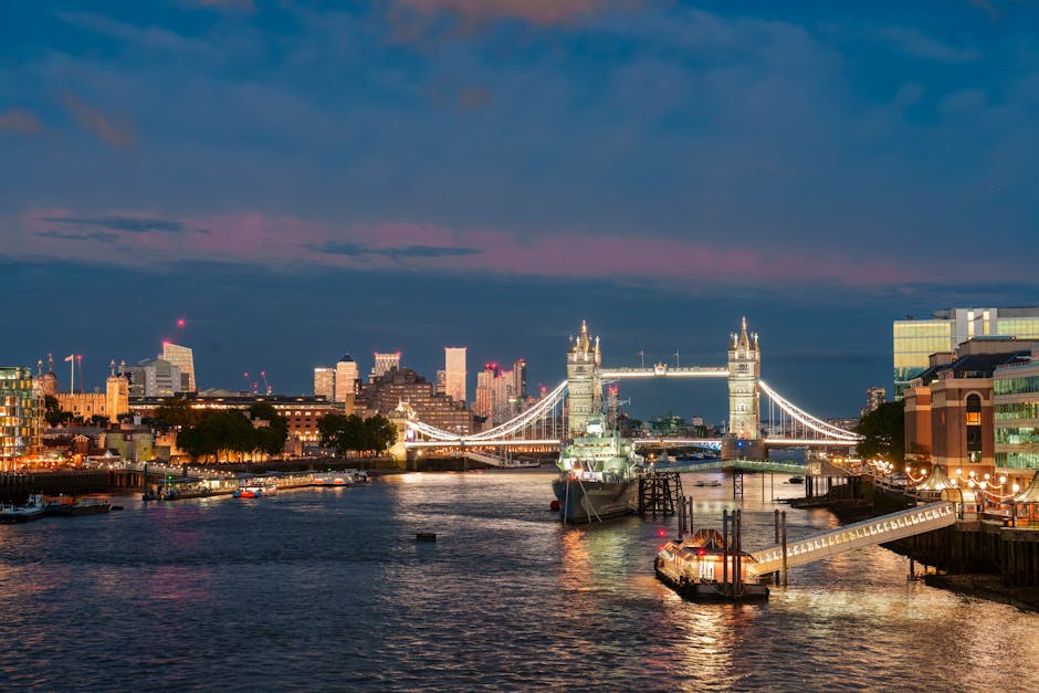 A panoramic view of the London skyline during twilight, featuring the illuminated Tower Bridge over the River Thames. On the left, modern office buildings and construction cranes are visible, while on the right, a mix of contemporary structures and residential areas are lined along the waterfront. The Thames reflects the city lights, with boats and vessels moored along the quayside. The bridge’s iconic towers and suspension cables are prominent against a darkening sky with subtle pink and purple hues. This scene illustrates an urban setting suitable for house removals, highlighting transport logistics and the importance of careful handling during home relocation or furniture transport in central London. The lighting emphasizes the quiet but busy atmosphere of the river area, and the overall scene supports content related to moving services, packing, and loading processes carried out in historic and modern parts of London, such as London Bridge and Borough.