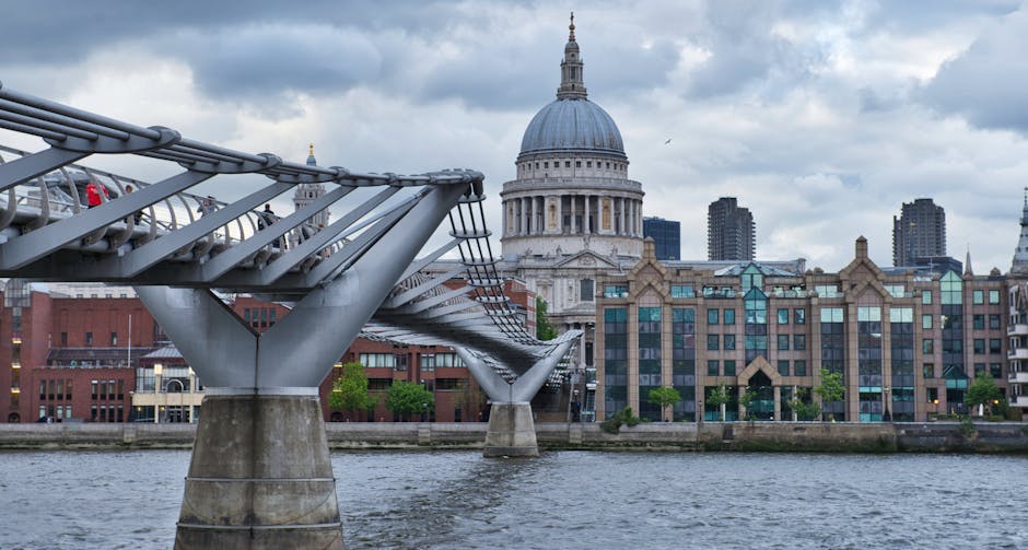 A panoramic view of the London skyline during twilight, featuring the illuminated Tower Bridge over the River Thames. On the left, modern office buildings and construction cranes are visible, while on the right, a mix of contemporary structures and residential areas are lined along the waterfront. The Thames reflects the city lights, with boats and vessels moored along the quayside. The bridge’s iconic towers and suspension cables are prominent against a darkening sky with subtle pink and purple hues. This scene illustrates an urban setting suitable for house removals, highlighting transport logistics and the importance of careful handling during home relocation or furniture transport in central London. The lighting emphasizes the quiet but busy atmosphere of the river area, and the overall scene supports content related to moving services, packing, and loading processes carried out in historic and modern parts of London, such as London Bridge and Borough.
