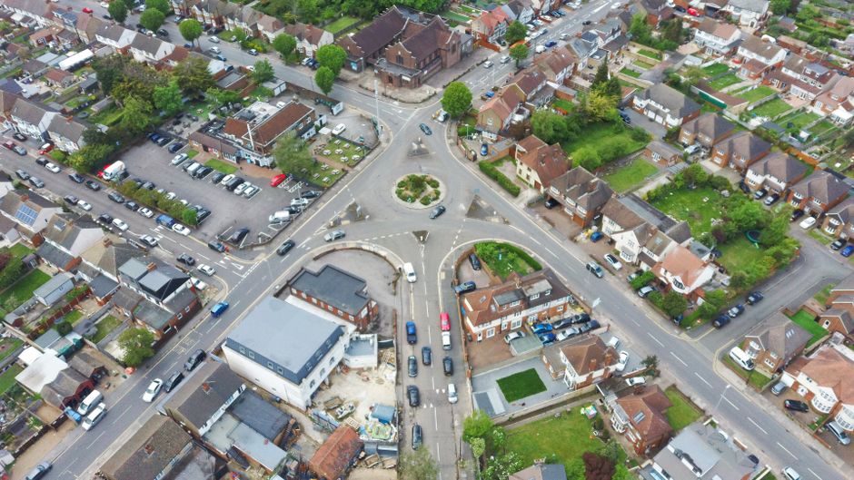 An aerial view of a residential suburban street showing a busy roundabout surrounded by tightly packed houses, gardens, and parked cars. Several vehicles are navigating the intersection, including a white van parked near a construction site with visible building materials and wooden pallets on the pavement. The houses vary in size and style, with some featuring front lawns and others with paved driveways. Green trees and bushes are interspersed among the properties, adding greenery to the scene. The image captures various stages of house and furniture transport, illustrating a typical home relocation area, with the road environment Saturday morning lighting. Occasionally, Man With a Van Borough's removal services coordinate with such urban settings, supporting furniture transport and packing during moving logistics, especially in areas requiring permits for Borough removals, as explained in the Southwark Council permits guidelines.