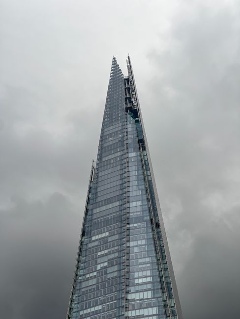 A tall, modern glass skyscraper with a sleek, angular design reaching high into an overcast sky, featuring reflective windows and visible construction or maintenance elements near the top. The building is part of the London skyline, with cloudy weather creating a muted background. The image emphasizes the building's height and architectural detail, relevant to urban property and relocation services provided by Man With a Van Borough, which specialises in house removals and furniture transport in Southwark. The building's reflective façade and towering presence highlight the scale of city-based relocations, fitting naturally into the context of home and business moving logistics in the borough.