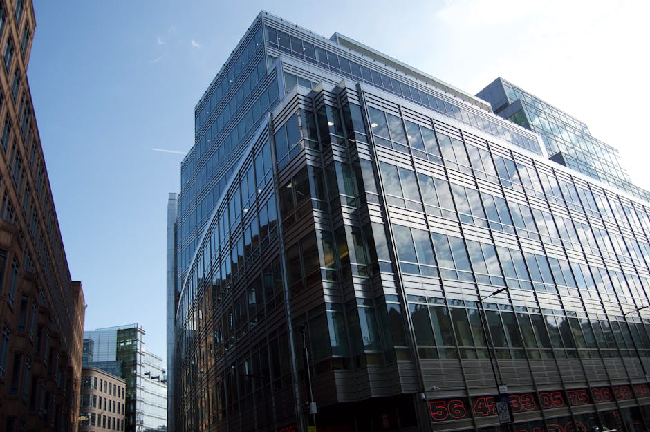 A modern multi-storey office building with a glass facade reflecting the blue sky and surrounding structures. The building's steel framework and large windows are visible, with some of the windows open. Adjacent to the office tower are other commercial buildings with brick and glass exteriors. The street level shows the pavement and parts of the building entrances. This image could depict an urban environment in central London, relevant to business relocations or office moves as offered by Man With a Van Borough, which specializes in house and office removals, including packing, transportation, and logistics for minimal downtime during relocations such as Tooley Street office moves.