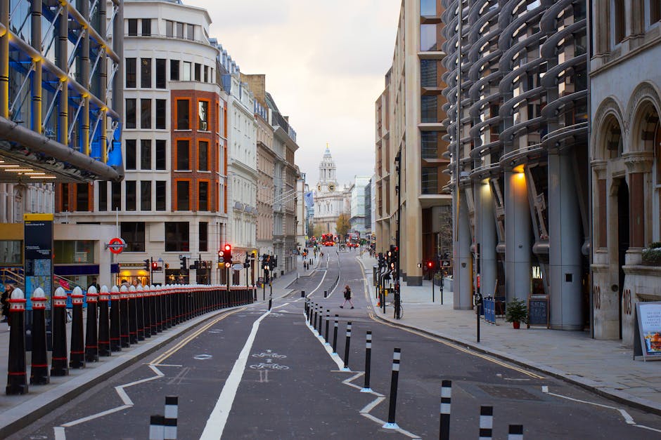 Photograph of an urban street scene featuring a narrow, paved roadway bordered by tall, modern office buildings and skyscrapers with glass and steel facades. On the left, a building with a brown stone exterior and arched windows, and on the right, a contemporary glass-fronted structure, frame the scene. Street signage and bollards are visible along the pavement, which is partly blocked by a small, green-leaved tree. The street appears quiet with minimal pedestrian activity, suggesting a typical city environment suitable for house or office relocations. The overcast sky provides diffuse natural lighting, and the scene reflects a professional setting where Man With a Van Borough might perform furniture transport or packing and moving services during urban property relocations, such as to or from Tooley Street offices.